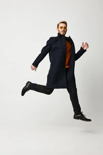 Un hombre guapo con un abrigo negro en pantalones y zapatos corre hacia un lado en pleno crecimiento sobre un fondo claro — Foto de Stock