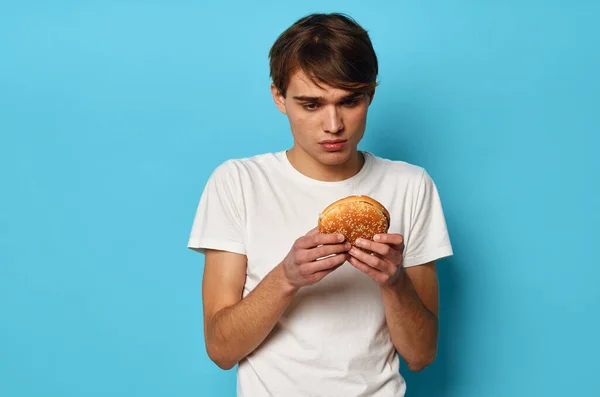흰색 티셔츠를 입고 햄버거를 손에 쥐고 파란 배경으로 점심 식사를 하는 남자 — 스톡 사진