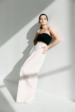 Siyah bluzlu ve beyaz pantolonlu bir kadın evin içinde duvara yaslanıyor.