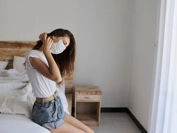 Žena na dovolené s lékařskou maskou na tváři sedí na posteli v izolovaném pokoji — Stock fotografie