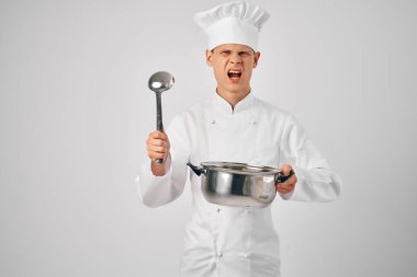 Elinde tava ve kepçeyle aşçı üniformalı bir adam yemek hazırlıyor.