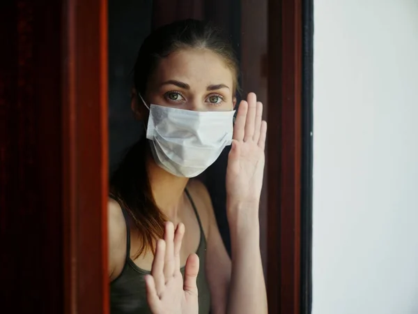 woman with sad facial expression in medical mask holds hands at lockdown window