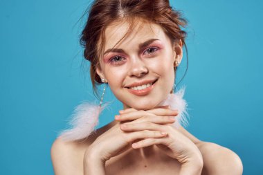 Çıplak omuzlu, tüylü küpeli, parlak, mavi makyajlı bir kadın.