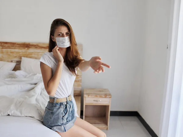 Jonge vrouw in een medisch masker zit op het bed en toont haar vinger naar de zijkant isolatie — Stockfoto