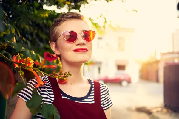 Ganska kort hår kvinna bär solglasögon buske med blommor utomhus — Stockfoto