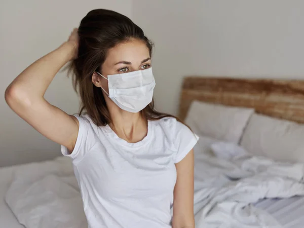 Woman in medical mask touches hair on her head with her hand and sits in bed interior — Stockfoto