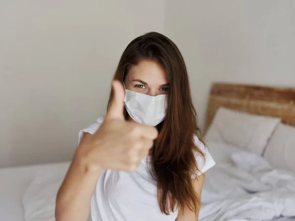 Woman sitting on bed in hotel thumb up medical mask — Stockfoto