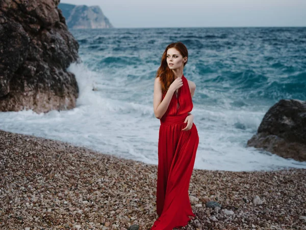 Όμορφη γυναίκα με κόκκινο φόρεμα ποζάρουν κοντά στο βράχο παραλία ρομαντική σχέση διακοπές — Φωτογραφία Αρχείου