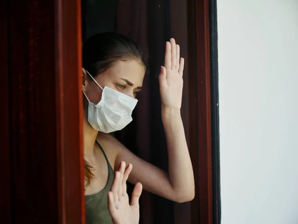 woman with sad facial expression in medical mask looking out the lockdown window