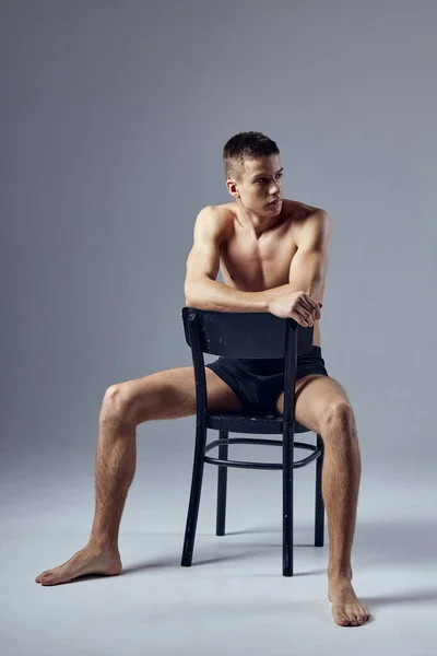 Cute guy black shorts sitting on a chair look to the side studio — Fotografia de Stock