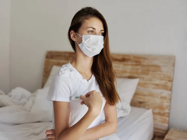 Woman in medical mask with a thermometer under her arm and sitting on the bed — Fotografia de Stock