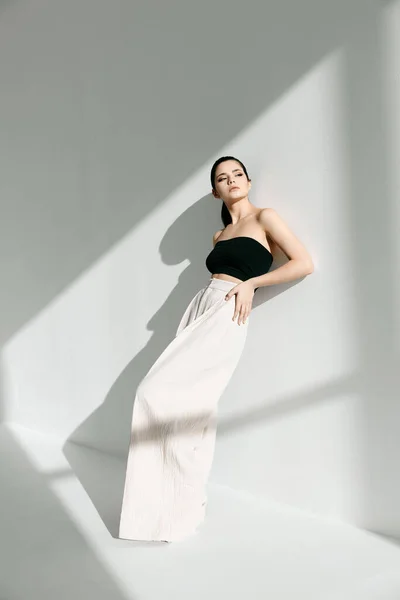 A fashionable woman in a black t-shirt and a white skirt is leaning against a wall indoors and a shadow is falling — Stock Photo, Image
