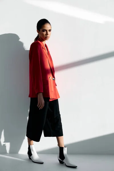 Uma mulher em uma jaqueta vermelha e calças da moda encostadas às botas de sapatos de outono de parede — Fotografia de Stock