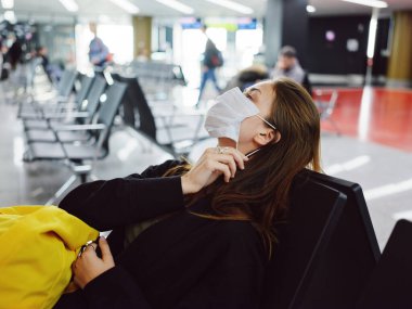 Tıbbi maske takan kadın başını arkaya eğmiş havaalanında oturmuş bekliyor.