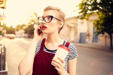 Gözlüklü kadın sokakta telefonla konuşuyor. Bir fincan kahve.