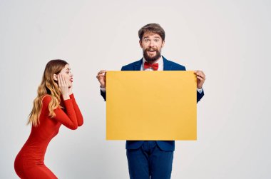 Takım elbiseli bir iş adamı kırmızı elbiseli bir kadının yanında sarı model posterler asıyor.