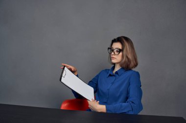 Mavi gömlekli bir kadın masada oturuyor. Elinde bir klasör var.