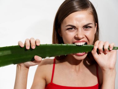 Dişlerinin arasında yeşil bir aloe yaprağı ve kırmızı bir tişört ve temiz bir cilt modeli tutan bir kadın.