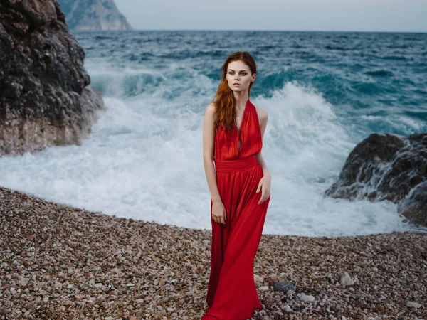 Ελκυστική γυναίκα σε κόκκινο φόρεμα από τους βράχους του ωκεανού ποζάρουν — Φωτογραφία Αρχείου