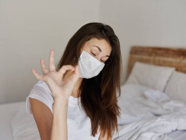 Yatak odasında eli tıbbi maskeli bir kadın.