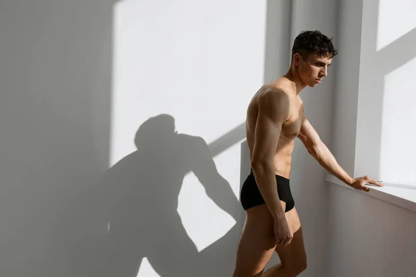Seorang pria dengan celana pendek hitam dekat jendela dengan latar belakang cahaya tubuh yang dipompa — Stok Foto