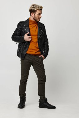 Turuncu kazaklı ve pantolonlu şık bir adam elini cebinden tutup yan tarafa bakıyor.