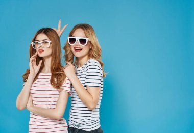 Çizgili tişörtlü iki tatlı kız arkadaş sarılır eğlence gözlükleri mavi arka planda