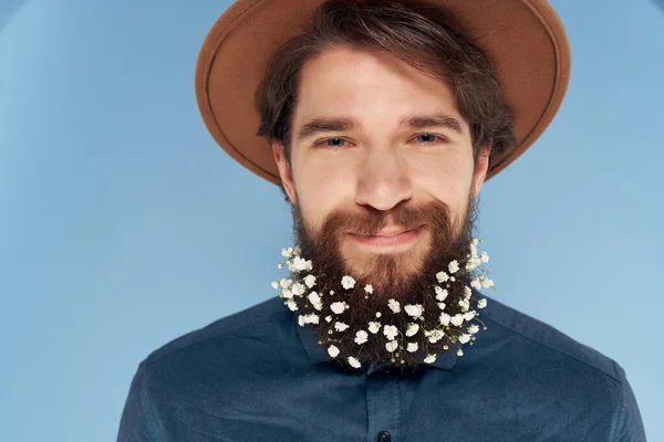 Homem alegre com flores em uma barba em um chapéu close-up fundo azul — Fotografia de Stock