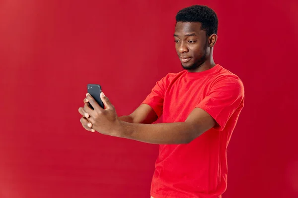 Man av afrikanskt utseende i en röd t-shirt med en telefon i händerna isolerad bakgrund — Stockfoto