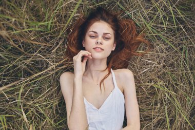 Beyaz elbiseli neşeli kızıl saçlı kadın çimenlerin üzerinde uzanıp dinleniyor.