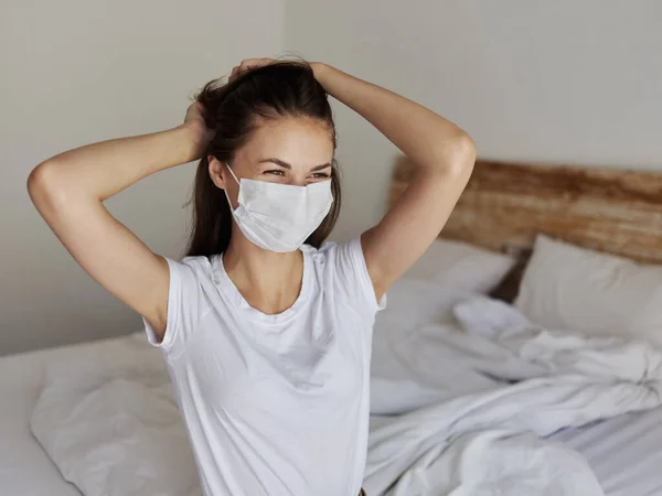 Eine Frau in medizinischer Maske sitzt auf einem Bett in einem hellen Raum und berührt ihren Kopf mit den Händen — Stockfoto