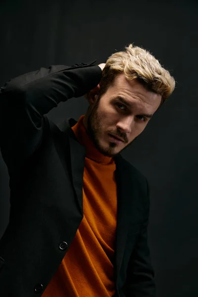 blond guy in an orange sweater and jacket on a dark background holds his hand behind his head close-up cropped view