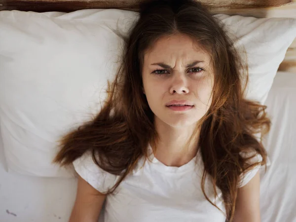 Mujer enojada yace en la cama en una camiseta blanca insatisfecha con un resfriado —  Fotos de Stock
