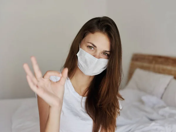 Frau mit medizinischer Maske sitzt auf Bett in Hotel positiver Handgeste — Stockfoto