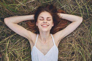 Beyaz elbiseli neşeli kızıl saçlı kadın çimenlerin üzerinde uzanıp dinleniyor.
