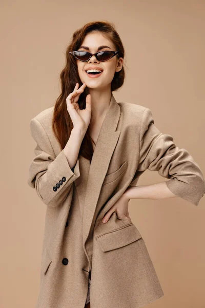 Happy woman in sunglasses and coat holds hand on waist on beige background — Stock Photo, Image