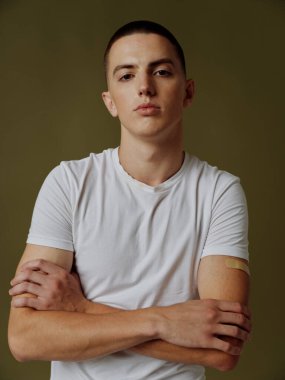 Beyaz tişörtlü bir adam kollarını koyu bir arka planda göğsünde kavuşturdu ve yapıştırıcı bir aşı pasaportu vardı.