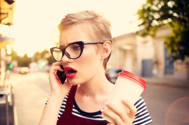 Gözlüklü kadın sokakta telefonla konuşuyor. Bir fincan kahve.