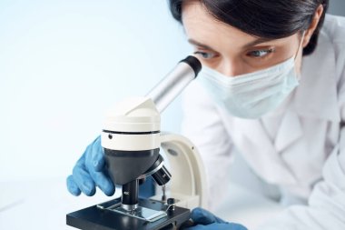 Tıbbi maskeli bir kadın laboratuvar asistanı mikroskop araştırmasına bakıyor.