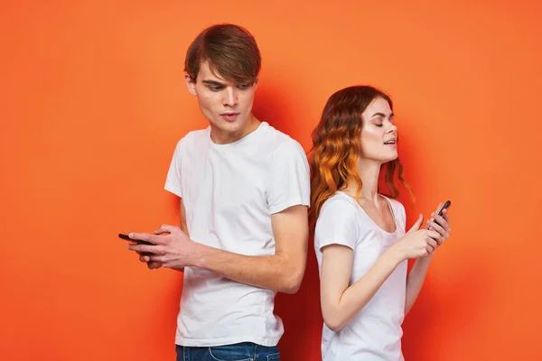 Fröhliches junges Paar in weißen T-Shirts Handys Unterhaltungstechnologie orange Hintergrund — Stockfoto