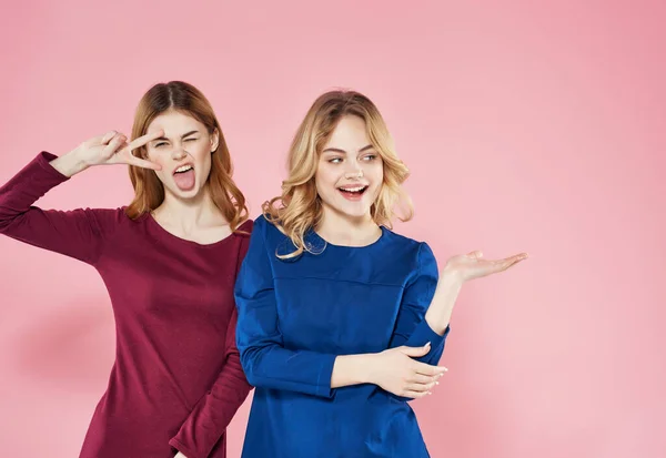 Dos mujeres alegres en vestidos están de pie junto a la amistad emociones fondo rosa —  Fotos de Stock