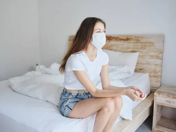 Vista laterale di bella donna in maschera medica seduta sul letto in camera isolata — Foto Stock