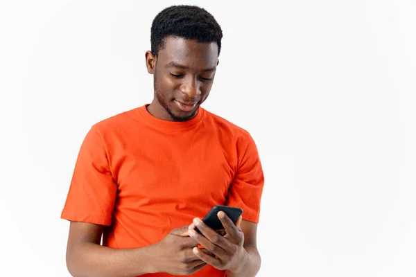 Um cara em uma camiseta laranja olha para a tela de um telefone celular em um fundo claro — Fotografia de Stock