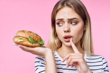 Çizgili tişörtlü, hamburgerli, lezzetli sandviçli pembe arka planlı kadın.