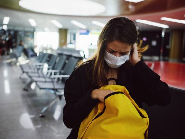 Sarı sırt çantalı, tıbbi maskeli yorgun kadın uçuşu bekliyor.