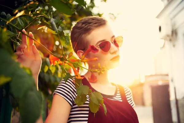Ganska kort hår kvinna bär solglasögon buske med blommor utomhus — Stockfoto