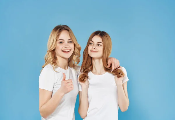 Two cute girlfriends in white t-shirts are standing next to friendship emotions blue background — Stock Photo, Image