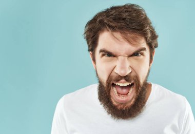 Beyaz tişörtlü sakallı bir adam öfke dolu bir el hareketi yapıyor.