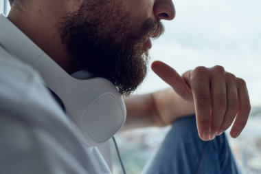 Beyaz tişörtlü bir adam pencerenin yanında oturmuş müzik dinliyor.