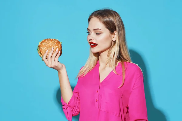 Allegra bionda in una camicia rosa hamburger fast food spuntino sfondo blu — Foto Stock